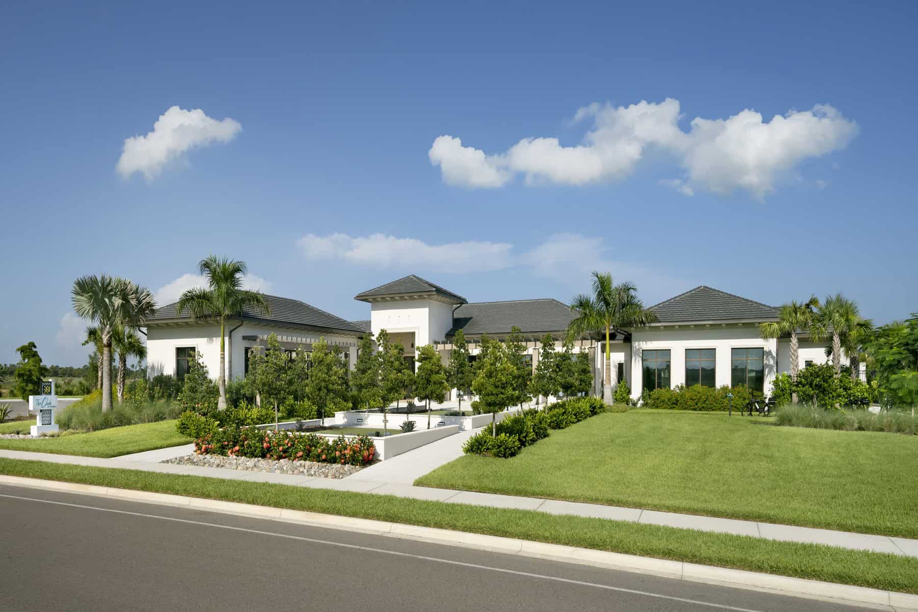 Exterior view of a single-story community clubhouse with a white stucco facade, dark tile roof, and central tower, framed by palm trees and a landscaped entry lawn.